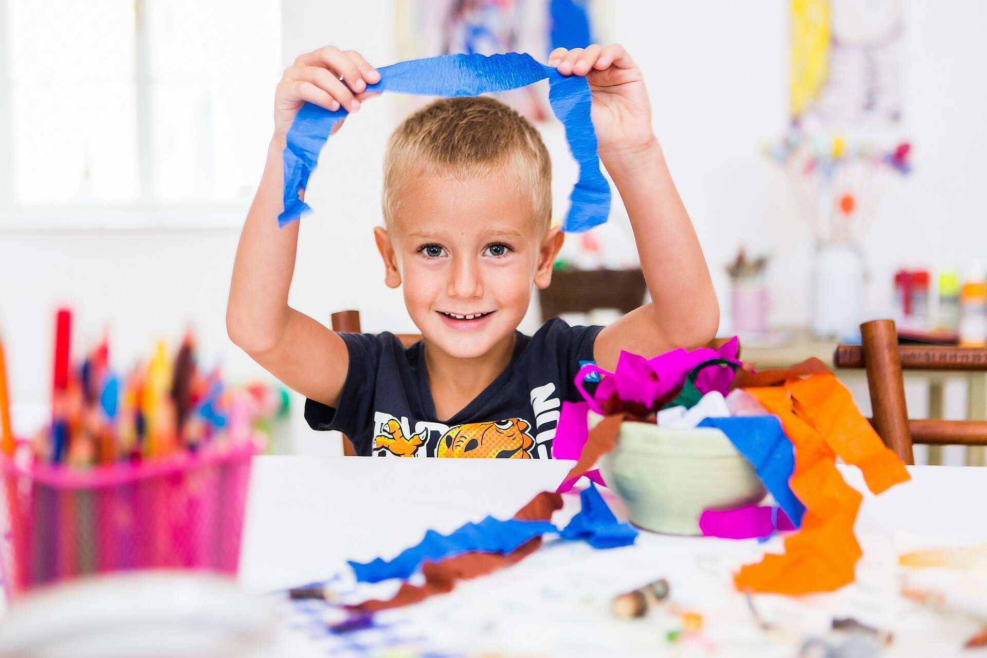 Kleiner blonder Junge hält ein Band in die Höhe, vor ihm liegen weitere bunte Bänder